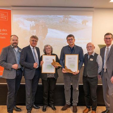 Mehrere Personen vor einer Bühne, zwei halten Urkunden in die Kamera